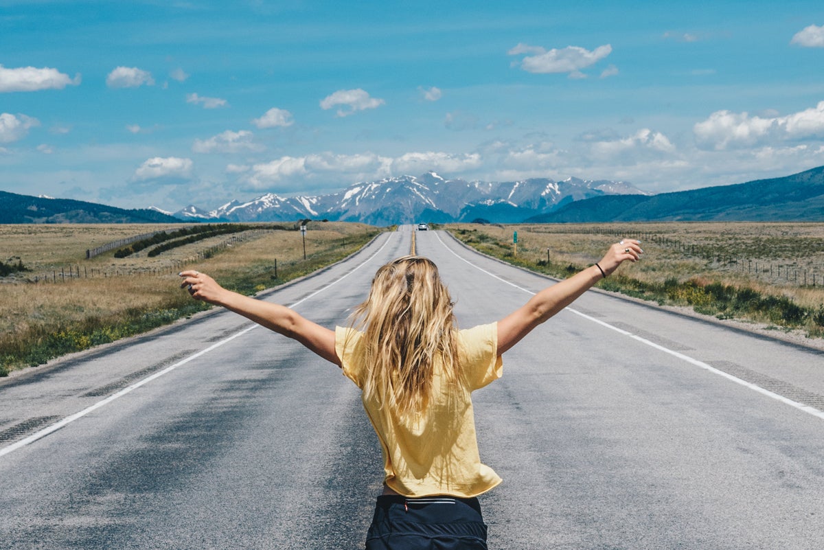 Girl with back to camera spread arms triumphantly in the middle of a one-lane highway creating a leading line to the mountains in the background