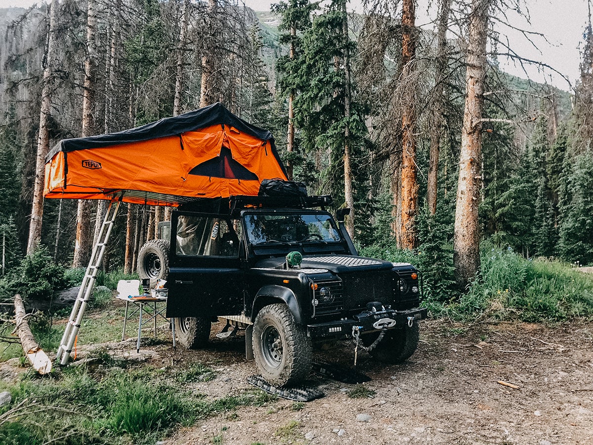 Old SUV surrounded by pine trees parked in dispersed mountain campsite with tent on top