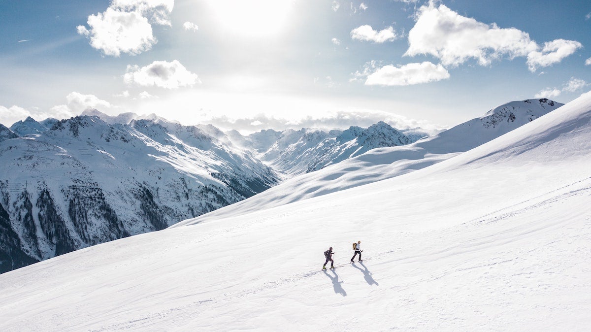 Two hikers working their way up a snowy mountain with peaks in the background