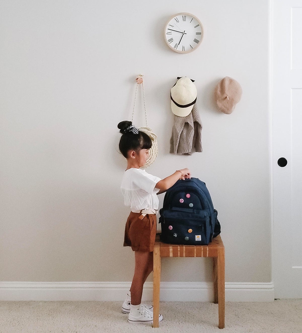Little girls zipping backpack in front of hats and bags hanging from wall coat hooks