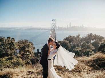 Photo of bride and groom kissing with bridge and city in background