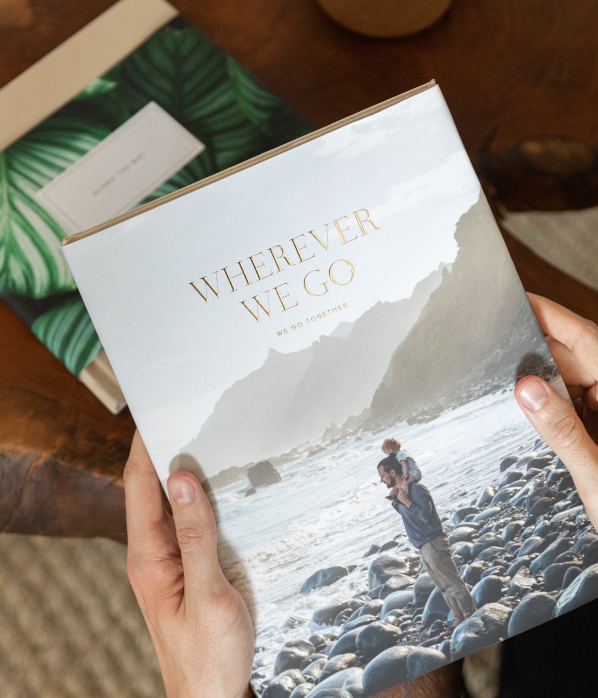 Hands holding up Artifact Uprising Hardcover Photo Book titled Wherever We Go featuring digital foil lettering and full dust jacket with photo of dad and baby on the beach