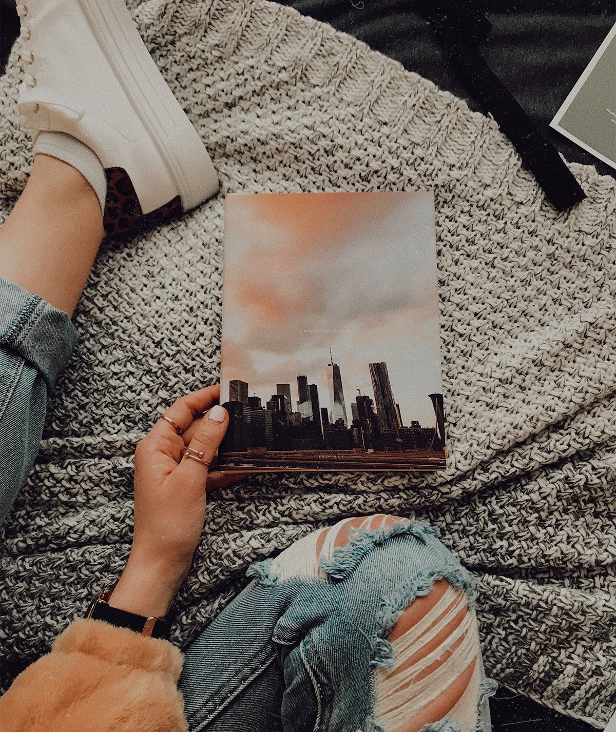 Woman sitting on bed with Artifact Uprising Hardcover Photo Book in her lap featuring photo of city skyline at sunset on the cover