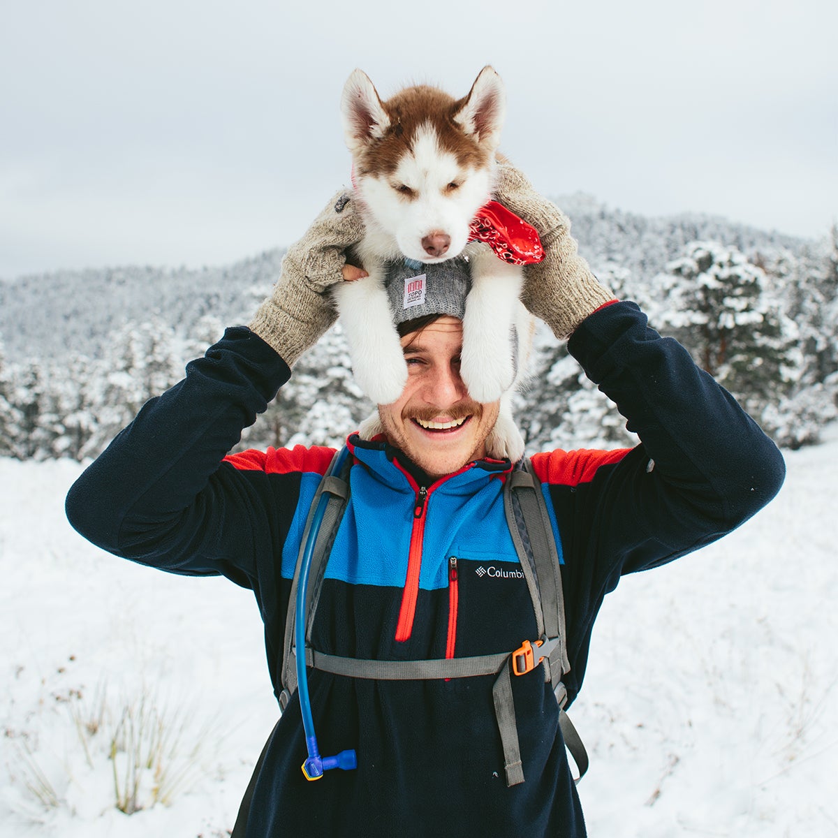 Man in snowy wilderness with husky puppy perched on his head
