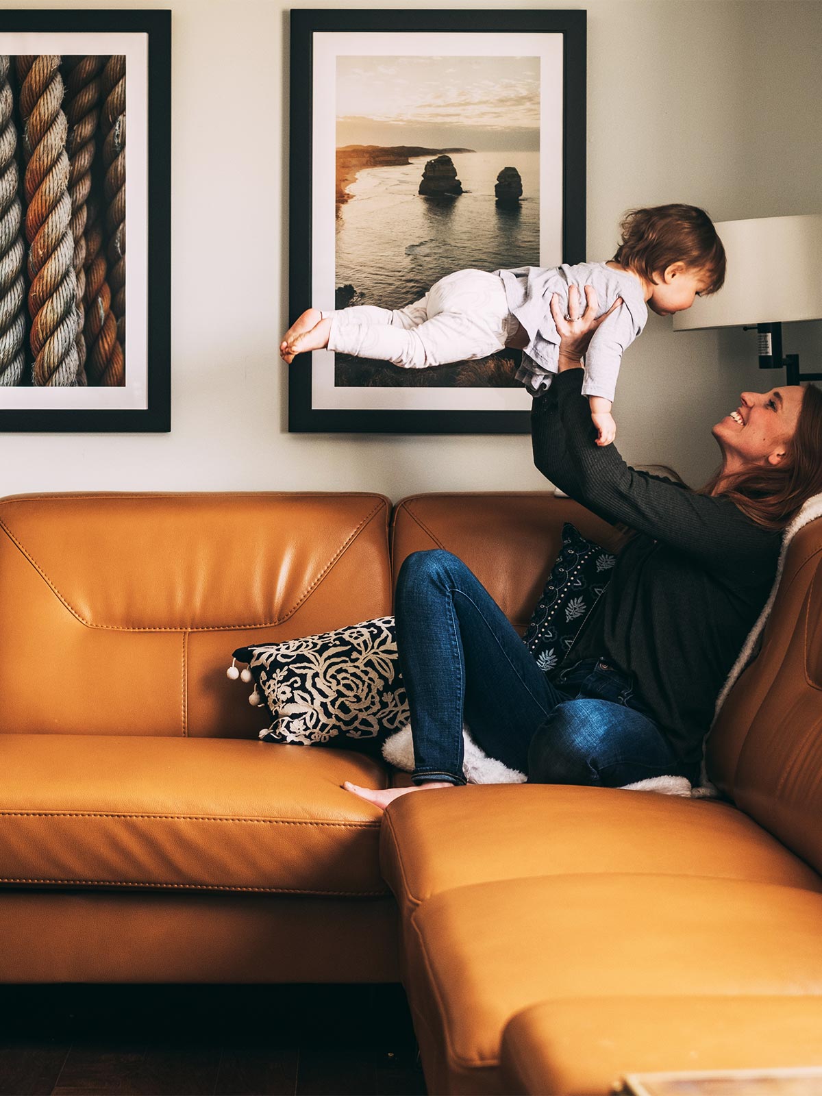 Mom holding toddler up in front of Gallery wall hanging above caramel leather couch