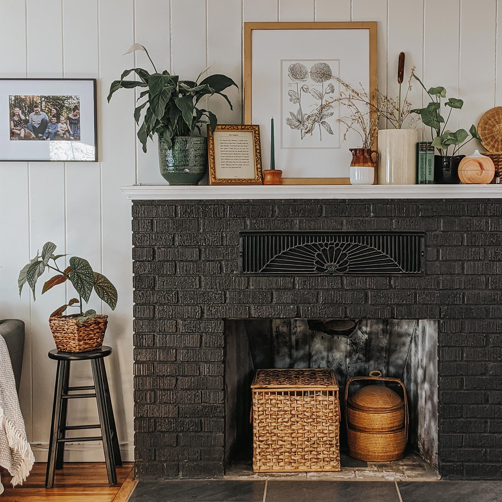 Mantel of black brick fireplace filled with frames, plants, and other ornamentals