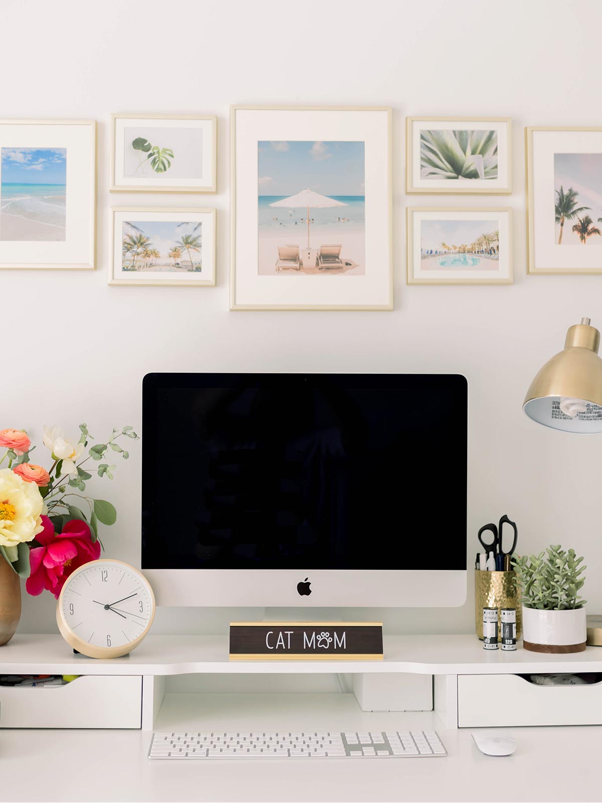 Gallery wall of frames above a desk with a computer, lamp, and bright bouquet.