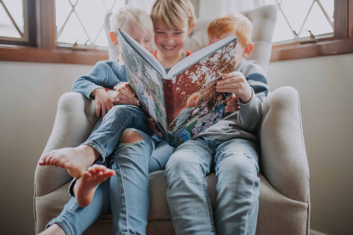 Three young siblings sitting in a big cream armchair flipping through an Artifact Uprising Hardcover Photo Book filled with family photos