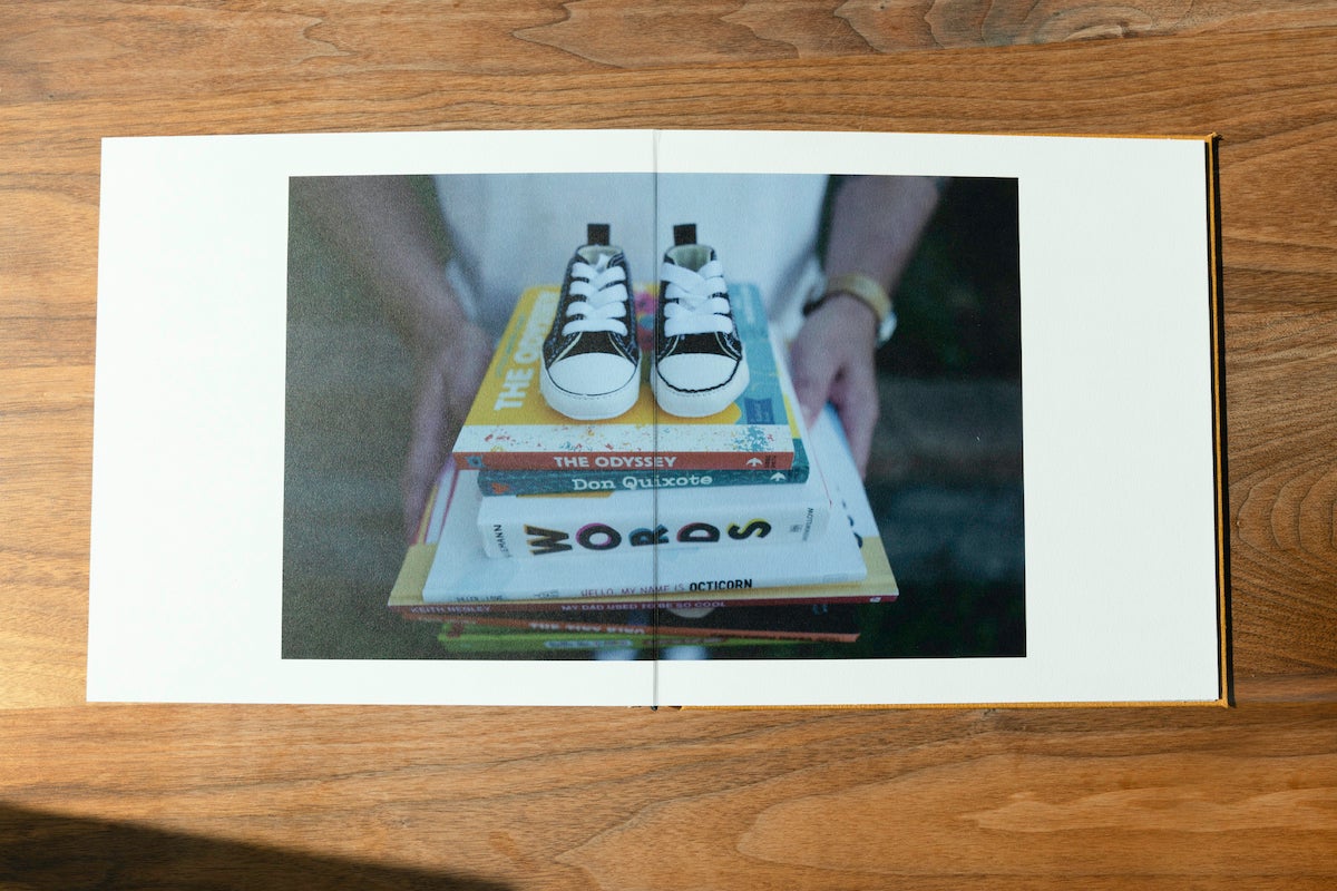 Artifact Uprising Layflat Photo Album opened to two page image of woman's hands holding stack of baby books with baby shoes on top