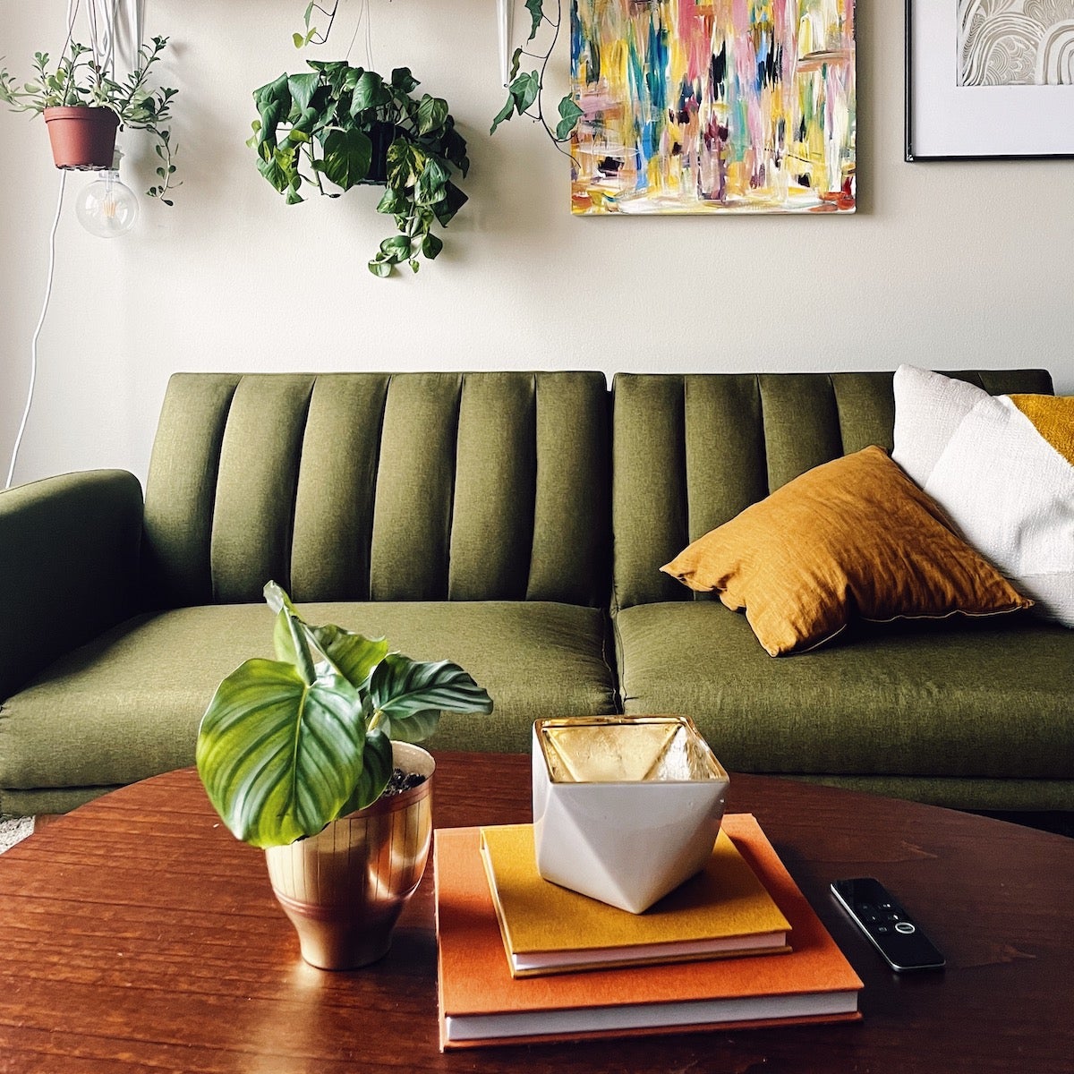 A colorful living with a green couch, yellow pillows, hanging plants and art, and then colorful coffee table books.