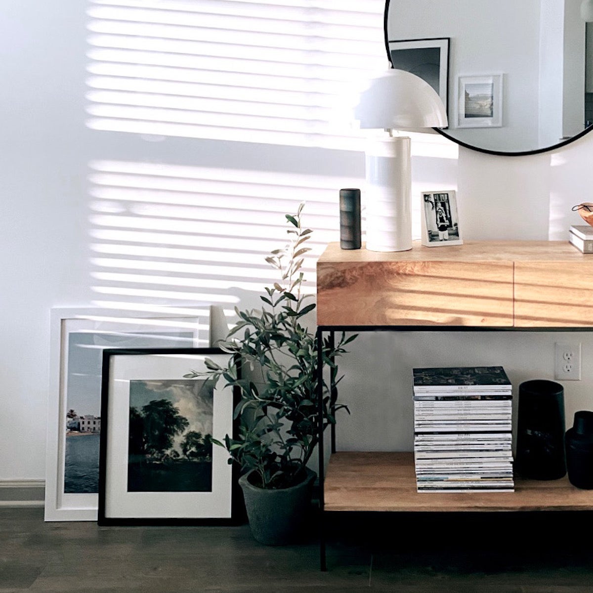 A console styled with books, lamps, and a decorative mirror — with a plant and two frames on the floor next to it. 
