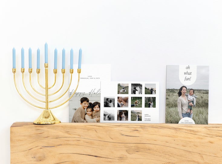 Three Artifact Uprising holiday cards on a wooden fireplace mantel next to a gold menorah with blue candles