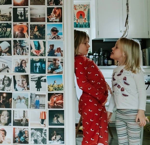 Two young girls leaning against a photo wall showing dozens of everyday prints of their family