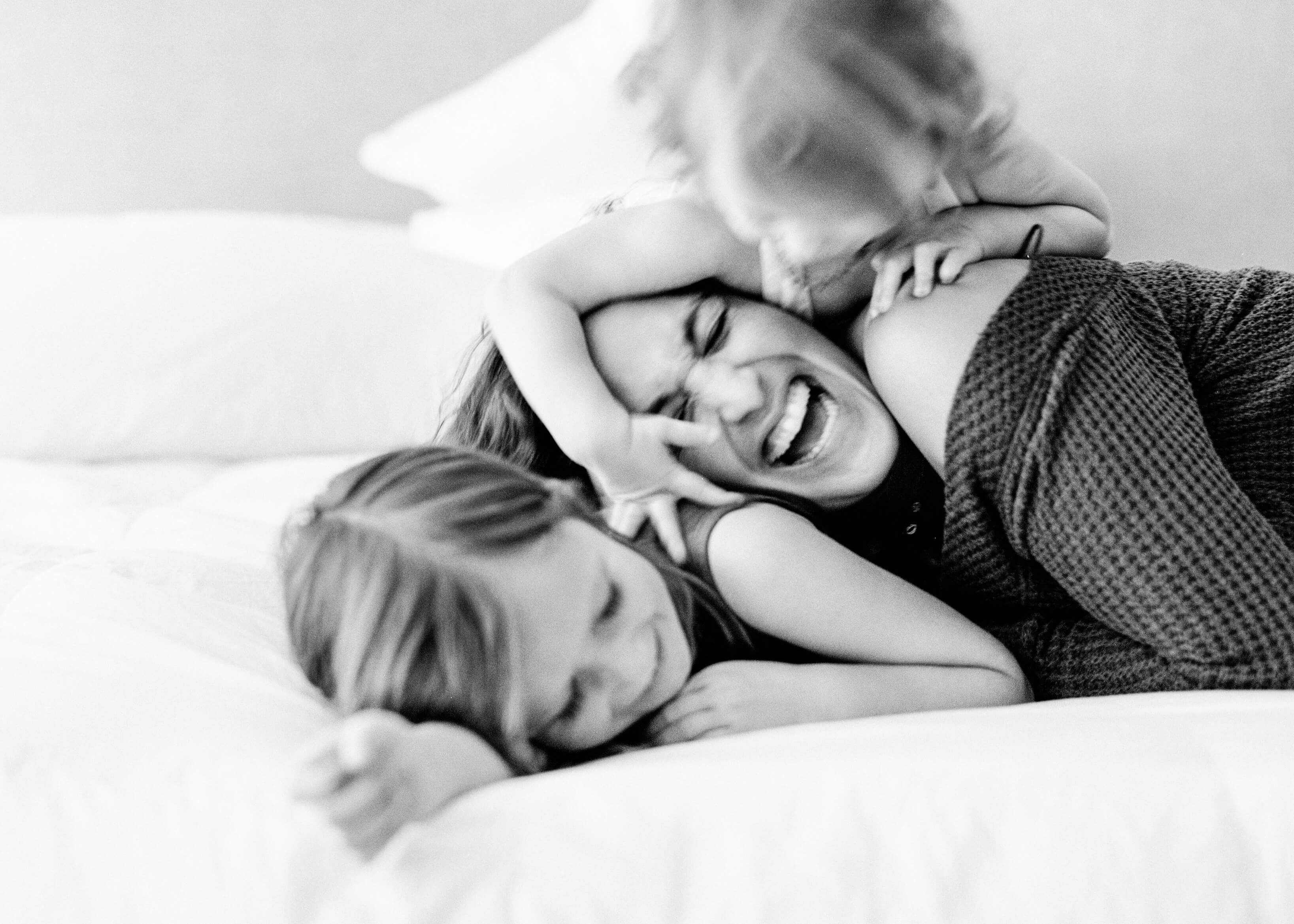 Little girl playfully jumping on top of mother and sister on bed
