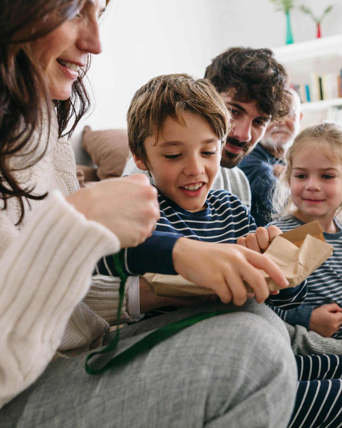 Boy smiling as he rips open gift wrapped in brown kraft paper on the couch with his family